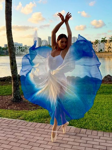 Ballerina in white leotard and blue and white long skirt dancing in front of a lake.