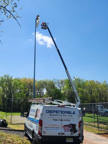 Workers repair stadium lighting using a crane and a van outdoors.