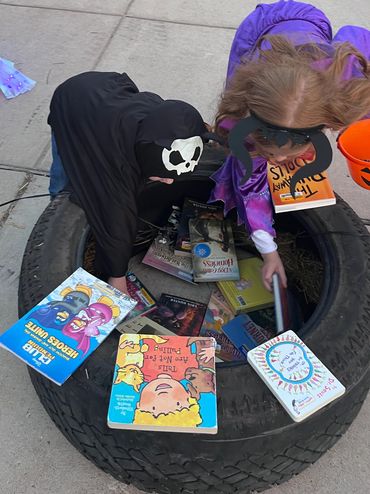 Children dressed in Halloween costumes excitedly digging through a large tire filled with books, cho
