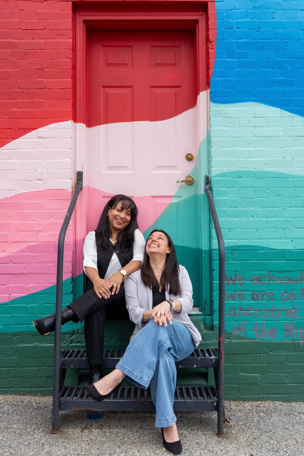 Two women sitting on steps with colorful painted door and wall behind them.
