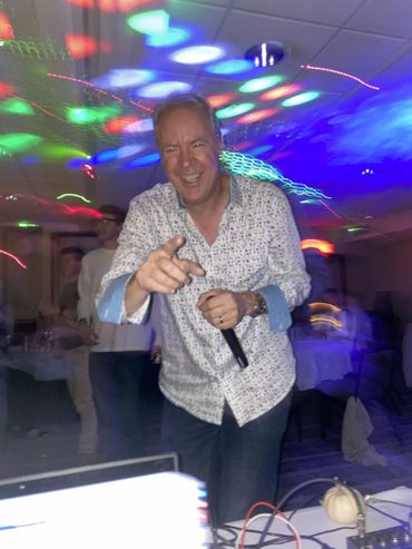 Man holding a microphone, smiling and pointing at a lively party with colorful lights.