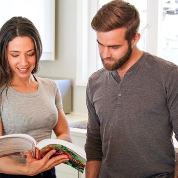 A couple reads a cookbook together in a bright kitchen.