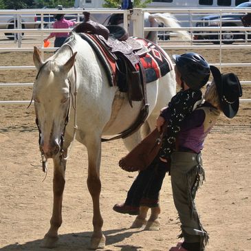 A young girl trying to lift another young rider up and help her get on a pony.