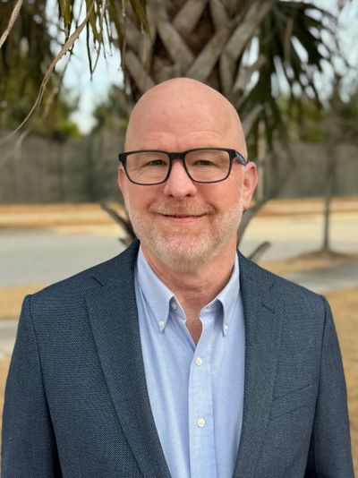 Smiling bald man with glasses in a blue blazer outdoors.