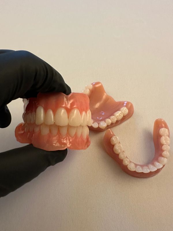 A gloved hand holding dentures with two sets of false teeth on a white surface.