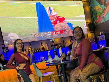 Two women in matching burgundy outfits watching a football game at a bar.