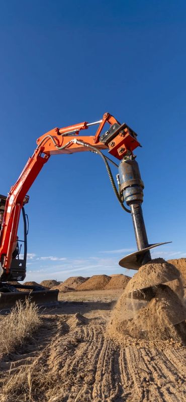 Excavator with auger attachment drilling into dry soil on a clear day.
