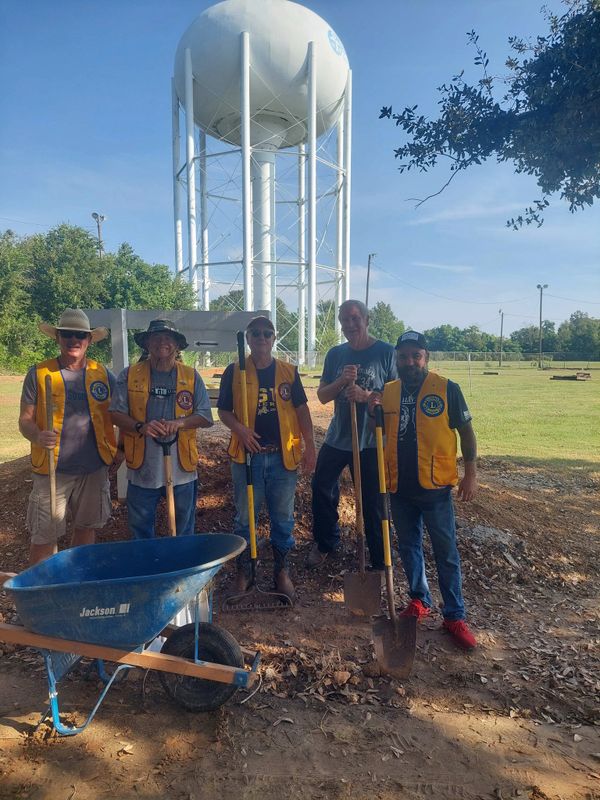 Five men in yellow vests with shovels and a wheelbarrow near a water tower.