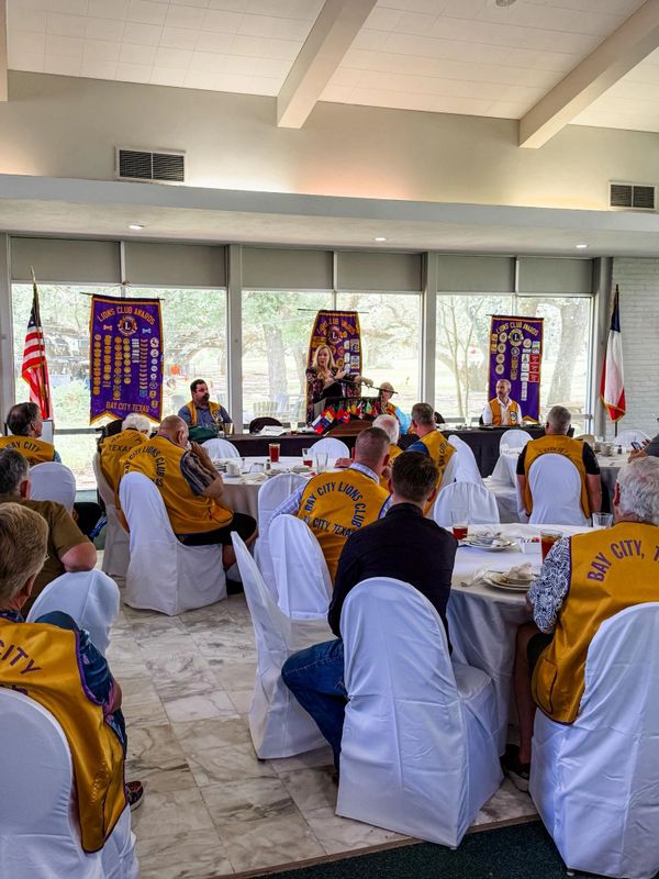 A Lions Club meeting with members in yellow vests seated and a woman speaking at the podium.