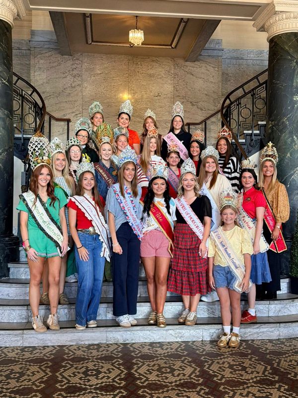 Group of young women and girls wearing crowns and sashes posing on stairs.
