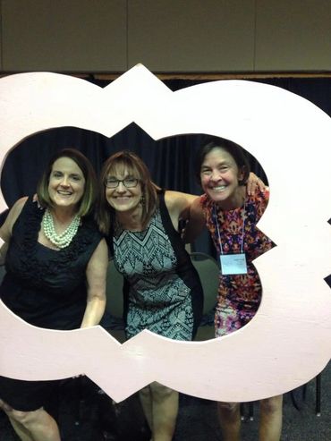 Three women smiling through a large decorative photo frame at an event.