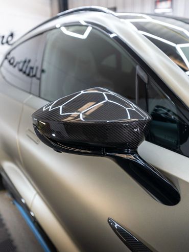Close-up of a car's carbon fiber side mirror on a matte silver vehicle.