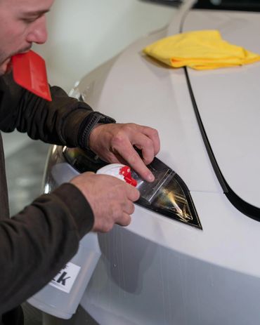 Man applying protective film on car headlight with spray bottle and red tool.