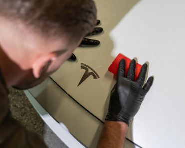 A person wearing black gloves applying a red tool near a Tesla logo on a white car.