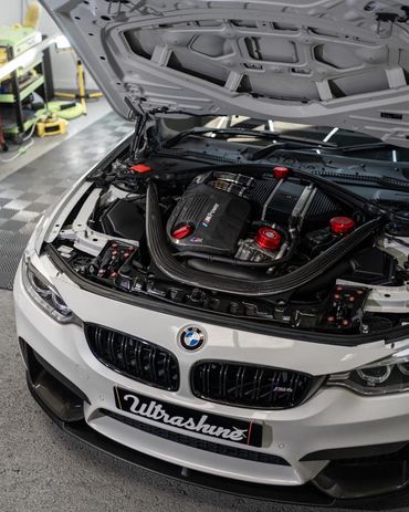 Open hood of a white BMW M4 showcasing a clean, modified engine bay with carbon fiber and red accents.
