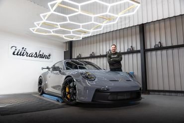 Man proudly stands next to a sleek gray Porsche in a modern garage.