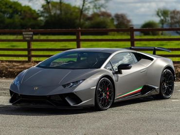 Sleek gray Lamborghini sports car parked on a sunny day with green fields in the background.
