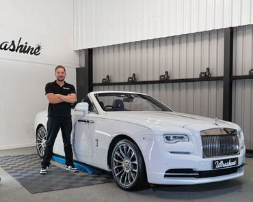Man stands proudly next to a shiny white Rolls-Royce convertible inside a garage.