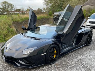 Black Lamborghini with scissor doors open, parked on gravel.