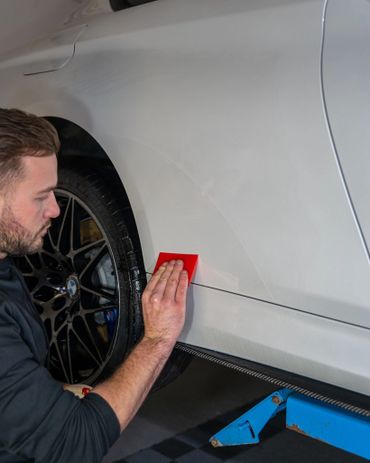 Man applying a red tool on a white car's side near the rear wheel.