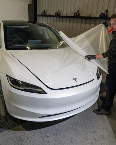 Person applying a protective film wrap on a white Tesla car hood indoors.