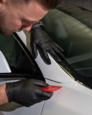 Person wearing black gloves applies film or tint to a car window with a red tool.