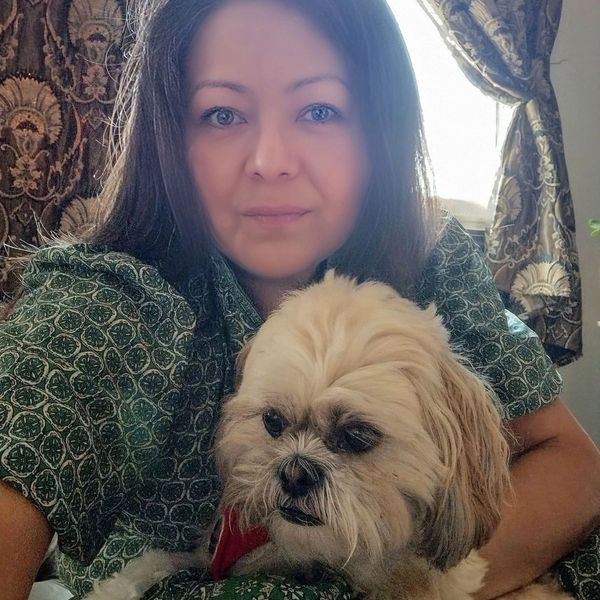 Woman with long hair poses with her small fluffy dog indoors near a window.