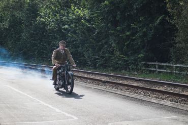 Man in vintage military uniform rides an old motorcycle beside train tracks.