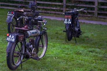 Three vintage motorcycles parked on grass near a wooden fence.