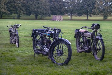 Three vintage motorcycles parked on a grassy field with trees in the background.