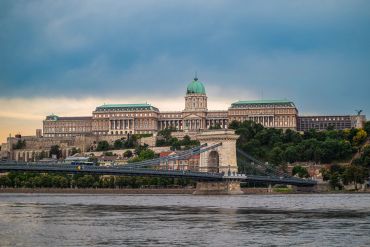 Buda Castle with a bridge over the Danube River under a cloudy sky.