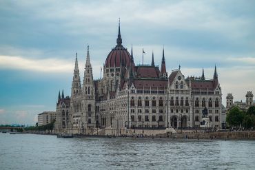 Historic building with spires beside a river under a cloudy sky.