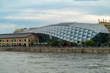 Modern glass dome blending with historic riverside building under a cloudy sky.