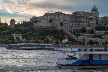 Boats sailing on a river with a historic castle in the background under a cloudy sky.