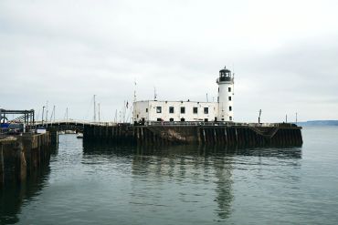 White lighthouse on a pier with calm water and boats in the background.