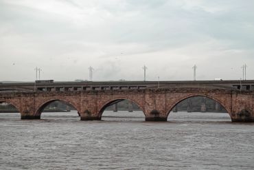 A train crosses a stone arch bridge over a river under a cloudy sky.