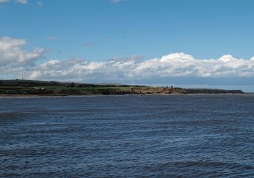 Coastal landscape with cliffs, green fields, and a large body of water under a blue sky.