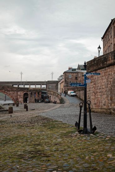 Cobblestone street with historic buildings and directional signs near a stone bridge.