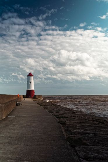 A red and white lighthouse at the end of a pier under a cloudy sky.