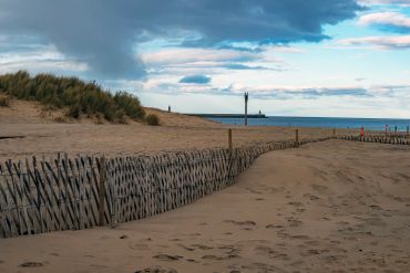 Sandy beach with wooden fences and cloudy sky near a lighthouse.