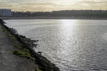 Calm water reflecting the cloudy sky near a rocky shoreline and distant buildings.