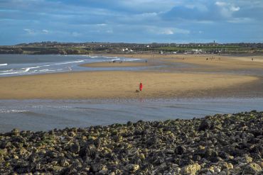 A lone person in red walks a dog on a vast sandy beach with rocky foreground and distant houses.