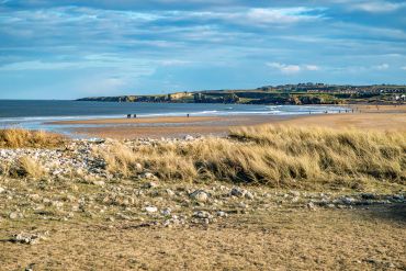 A wide sandy beach with people walking and a distant lighthouse under a cloudy sky.