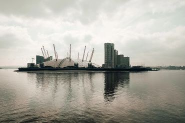 The O2 Arena in London with surrounding modern buildings reflected in the water on a cloudy day.