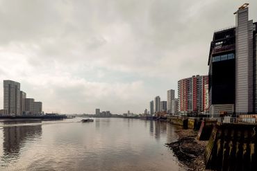 Boat cruising on a river with city skyline under cloudy sky.