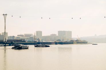 Cable cars glide over river with boats and city skyline in the background.