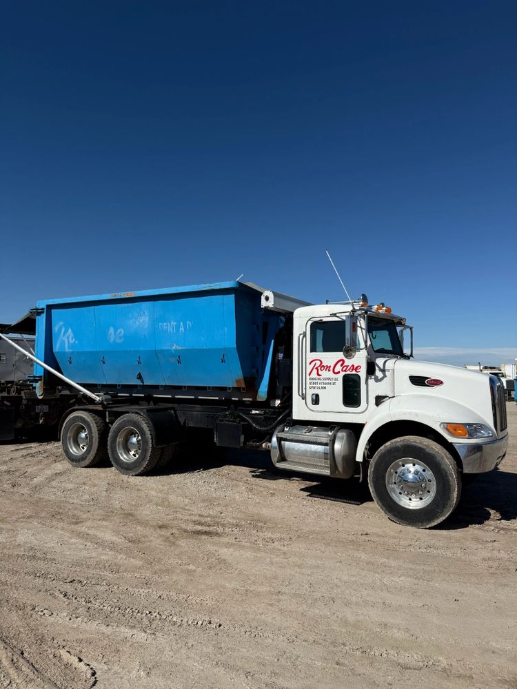 White and blue dump truck with Ron Case Roofing Supply logo on a dirt lot.