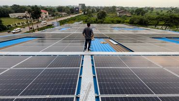 Man stands on rooftop covered with solar panels overlooking greenery and buildings.
