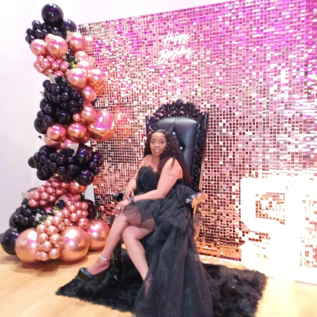 Woman in elegant black dress sits on a throne with balloon decorations.