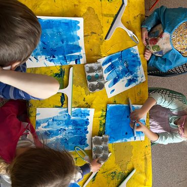 Children creating blue paintings at a yellow-covered table with various art supplies.